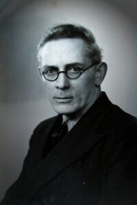 Black and white portrait photo of author Michael J. MacManus looking at the viewer, wearing a collared shirt and round-rimmed glasses.