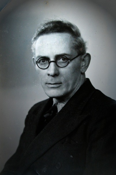 Black and white portrait photo of author Michael J. MacManus looking at the viewer, wearing a collared shirt and round-rimmed glasses.